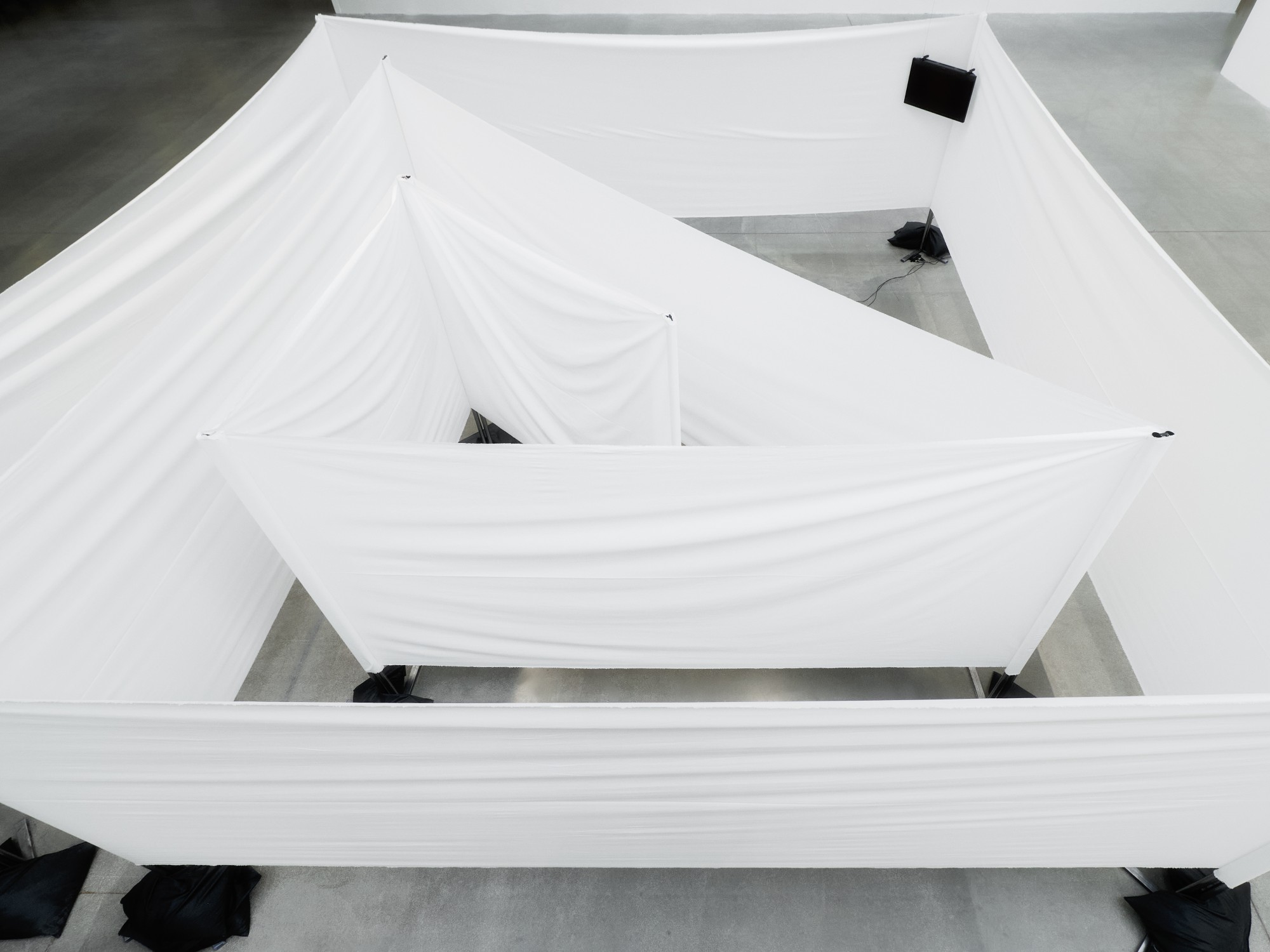 The labyrinth seen from above: nested walls of white fabric stretched between poles weighted with black sandbags.