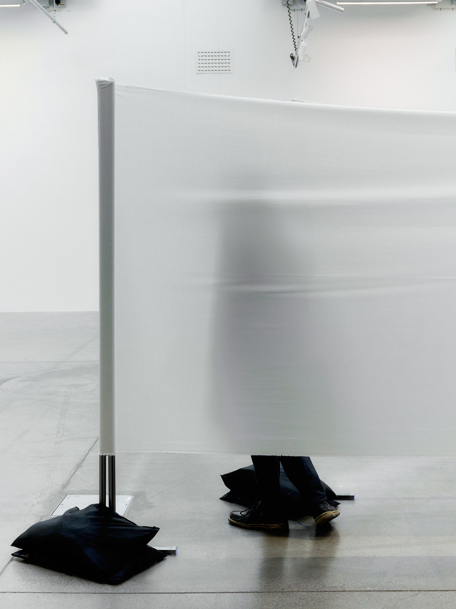 A person blurred behind a translucent wall of white fabric, their legs visible beneath it, next to a pole weighted with black sandbags.